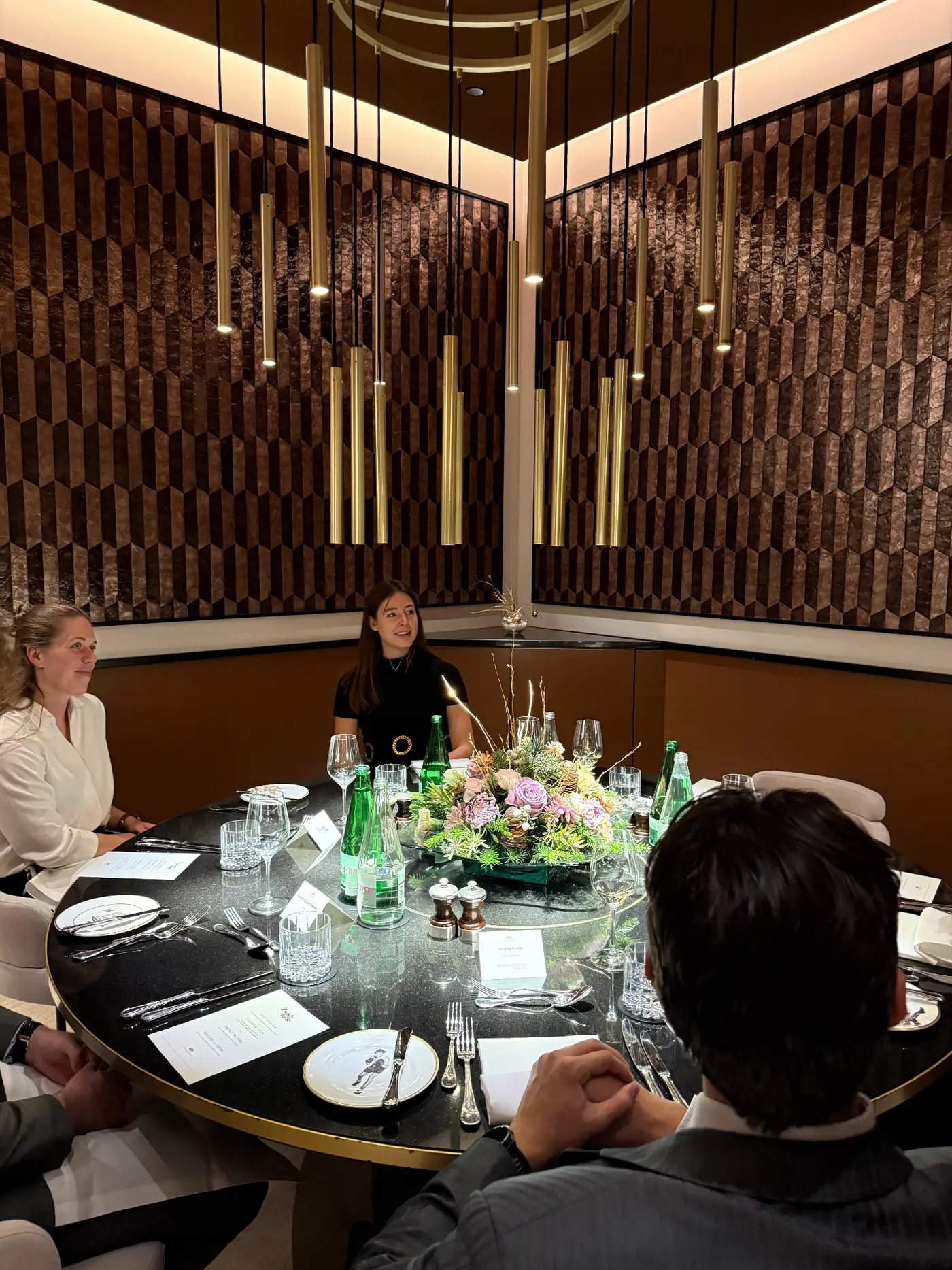 A Zurich Table dinner — guests around the round table beneath suspended brass pendants